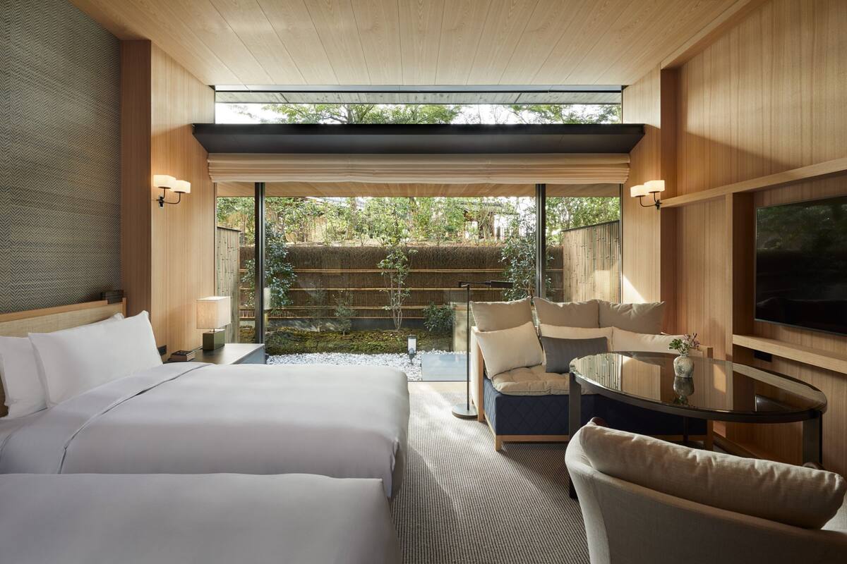Ein modernes Hotelzimmer mit einem großen Bett, einer Couch, einem Fernseher und Blick auf einen Garten durch große Fenster.