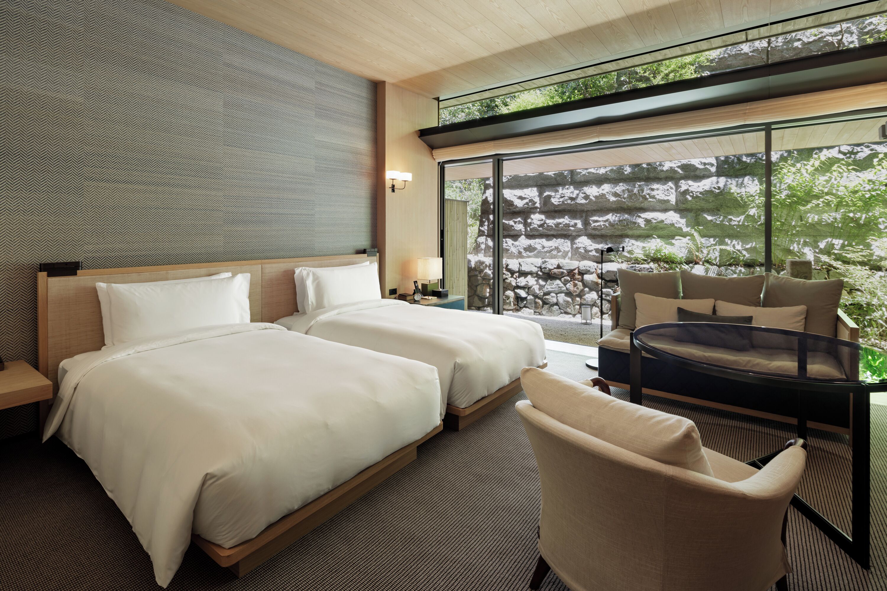 Quarto de hotel moderno com duas camas, um sofá, uma mesa de vidro e vista para um jardim rochoso.