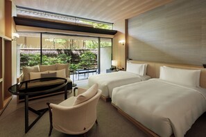 A hotel room with two beds, a seating area with a table and chairs, and a view of a garden through large windows.