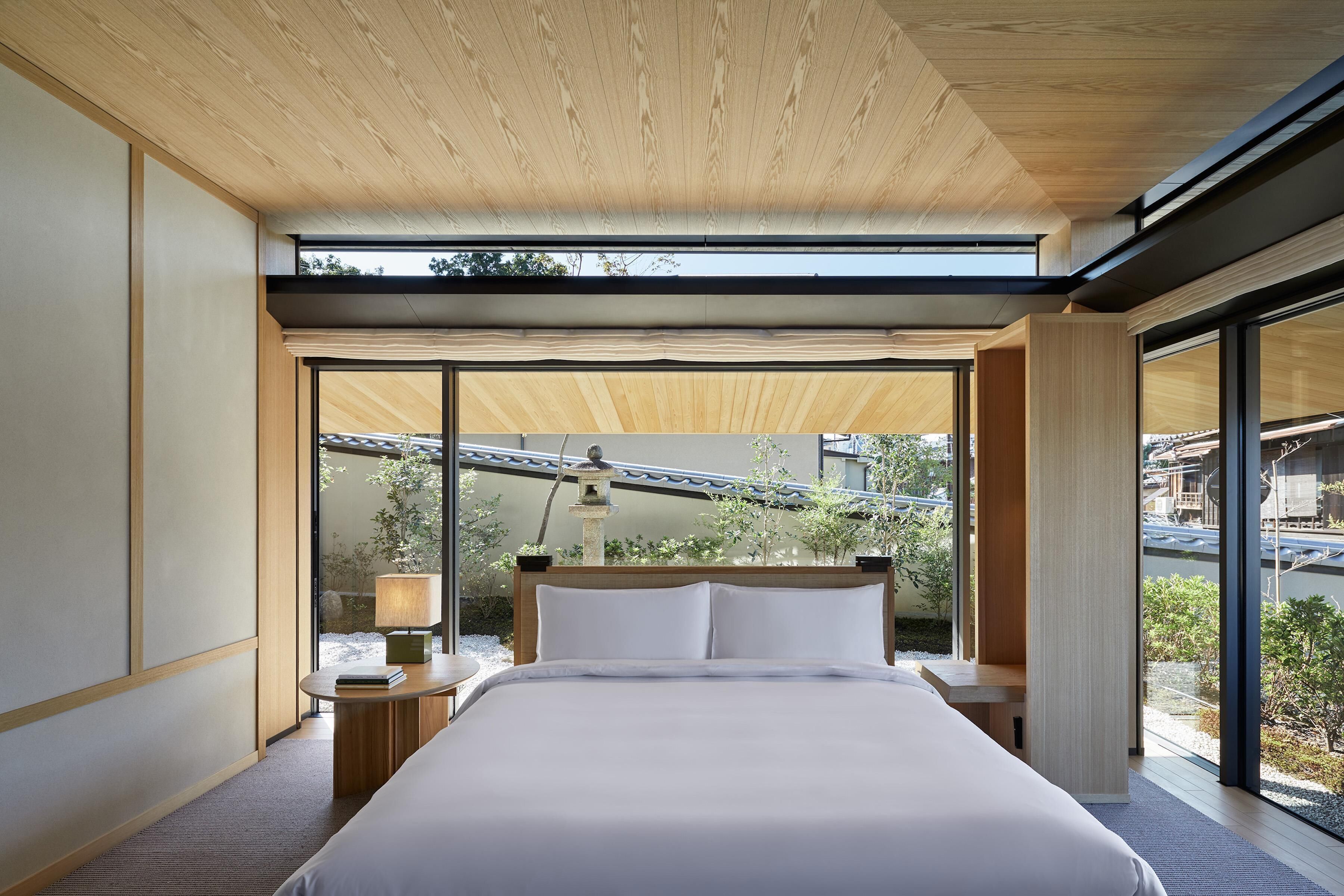 A modern bedroom with a large bed, a bedside table, a lamp, and a view of the outdoors through large windows.