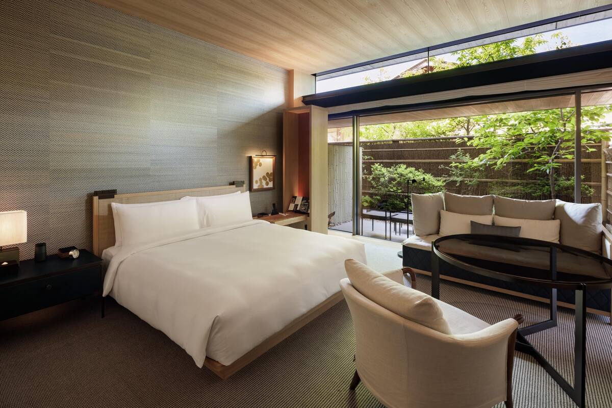 Ein modernes Hotelzimmer mit einem großen Bett, einer Sitzecke mit rundem Tisch und Blick auf einen Garten durch große Glasfenster.