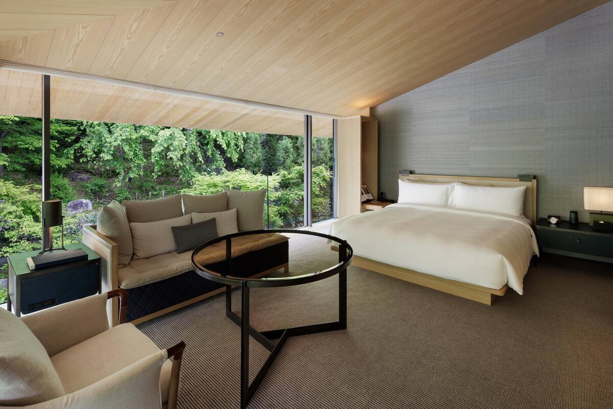Ein modernes Hotelzimmer mit einem großen Bett, einer Couch, einem Glastisch und Blick auf üppiges Grün.