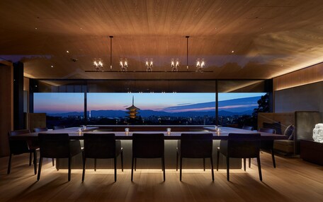 A modern dining area with a large table and chairs, a wooden ceiling, and a view of a cityscape at dusk.