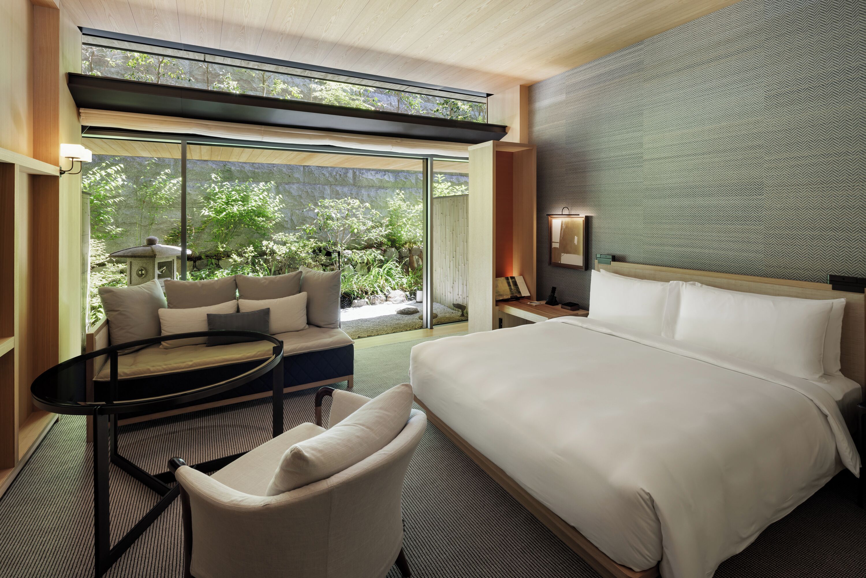 Une chambre d’hôtel moderne dotée d’un grand lit, d’un canapé, d’une petite table et offrant une vue sur un jardin grâce à de grandes fenêtres.