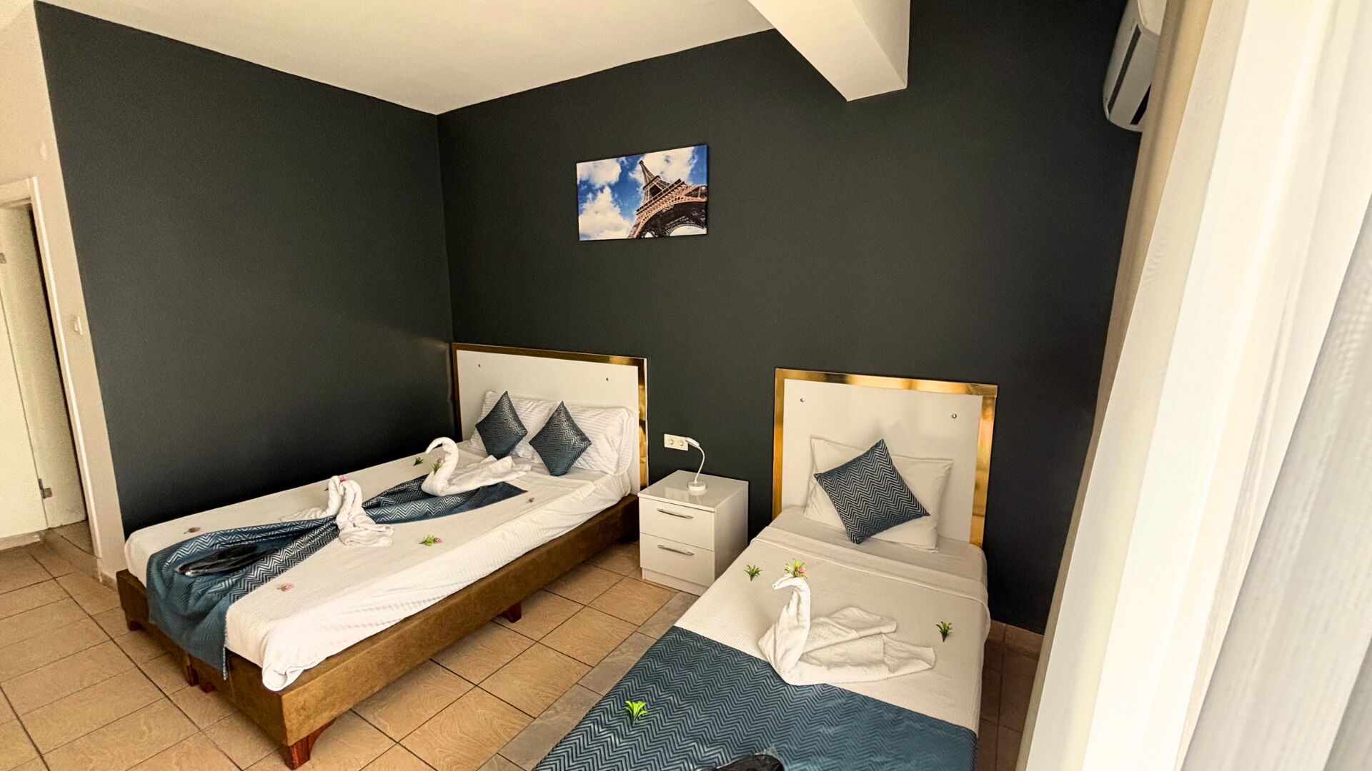 A hotel room with two beds, a small bedside table with a lamp, a wall-mounted picture of the Eiffel Tower, and a tiled floor.