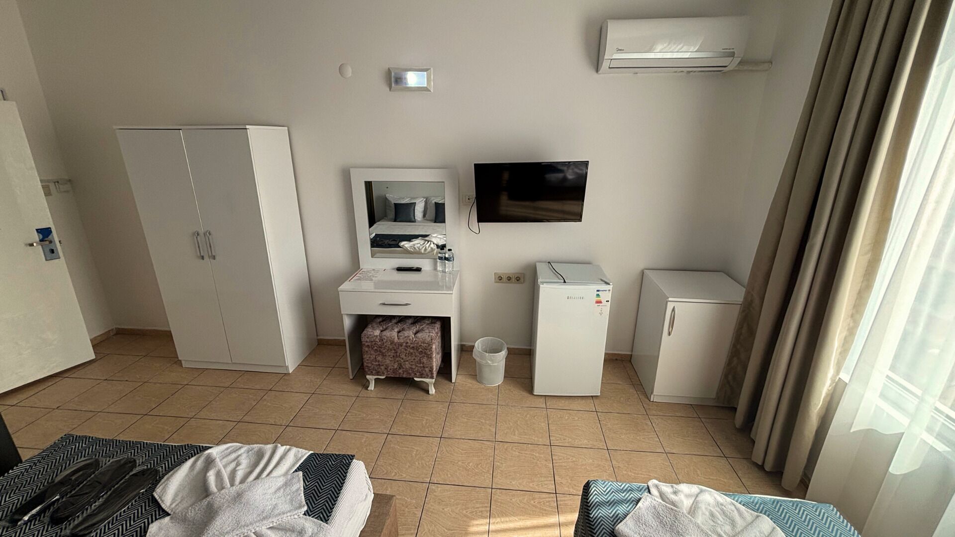 A hotel room with a bed, a desk with a mirror, a television, a refrigerator, and a wardrobe.