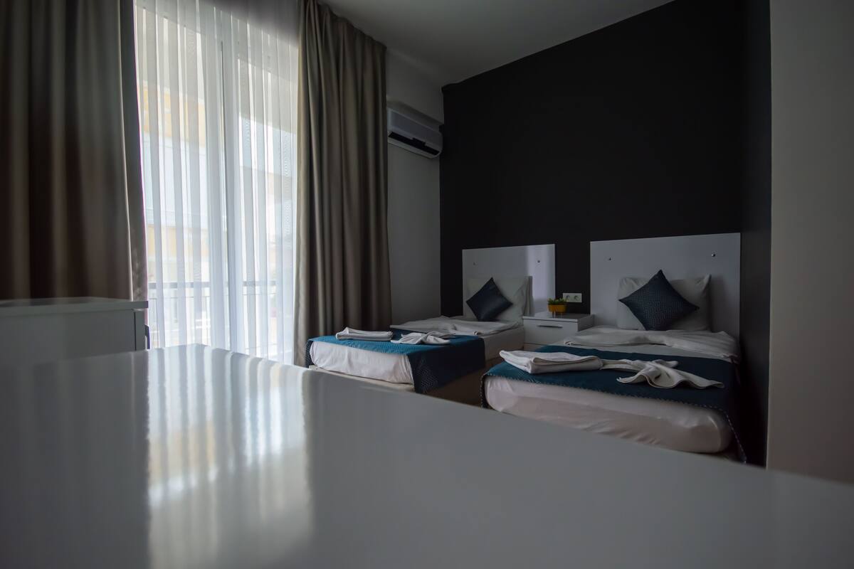 A hotel room with two beds, a large window with curtains, an air conditioning unit, and a white table.
