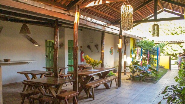 A covered outdoor dining area with wooden tables and benches, a tiled floor, and a hanging light fixture. - Pousada Vale Encantado (São Miguel dos Milagres)