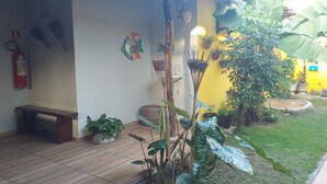 A patio area with a wooden bench, potted plants, a fire extinguisher, and a yellow wall with hanging decorations. - Pousada Vale Encantado (São Miguel dos Milagres)