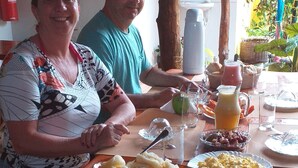 A dining table with various fruits and a pitcher of juice, surrounded by two people. - Pousada Vale Encantado (São Miguel dos Milagres)