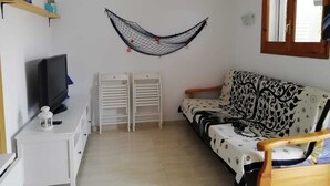 Una habitación pequeña y bien iluminada con un sofá, un televisor, un armario blanco y una hamaca.
