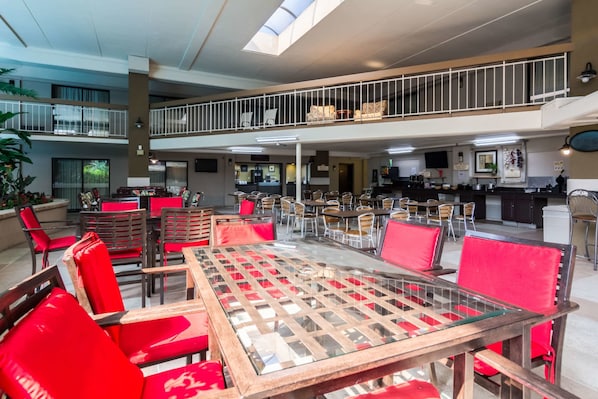 A spacious indoor area with multiple dining tables and chairs, a balcony with railings, and a kitchen area in the background.
