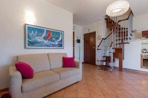 Un soggiorno con un divano color beige, un quadro con barche a vela, una scala e una zona cucina.
