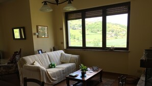 Een gezellige woonkamer met een bank, een bijzettafeltje en uitzicht op een groen landschap door grote ramen.