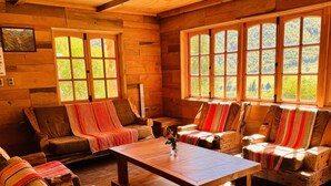 A cozy cabin interior with wooden walls, large windows, and a central wooden table surrounded by cushioned seating.
