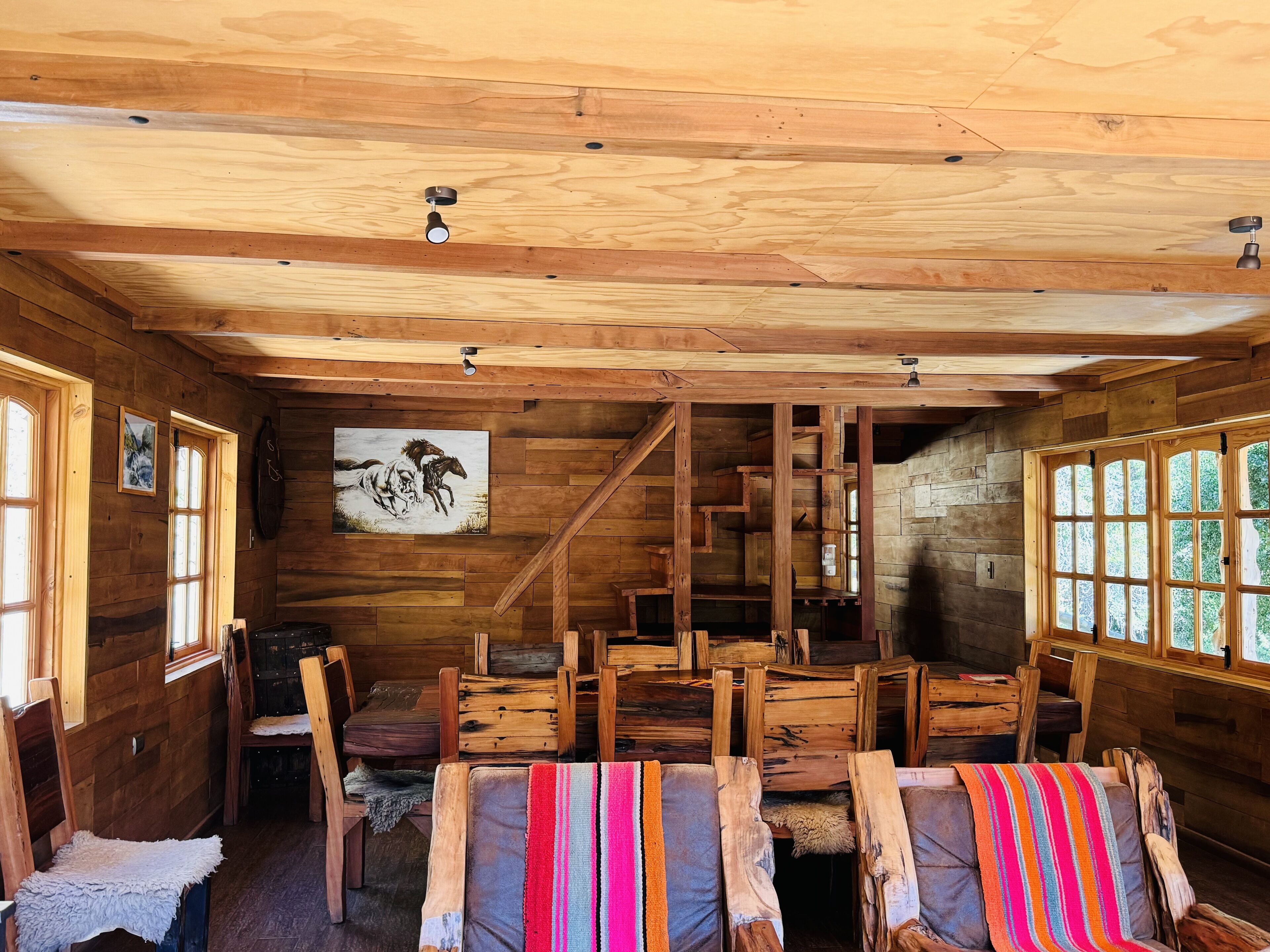 A rustic wooden cabin interior with a dining area, wooden chairs, a staircase, and a painting on the wall.