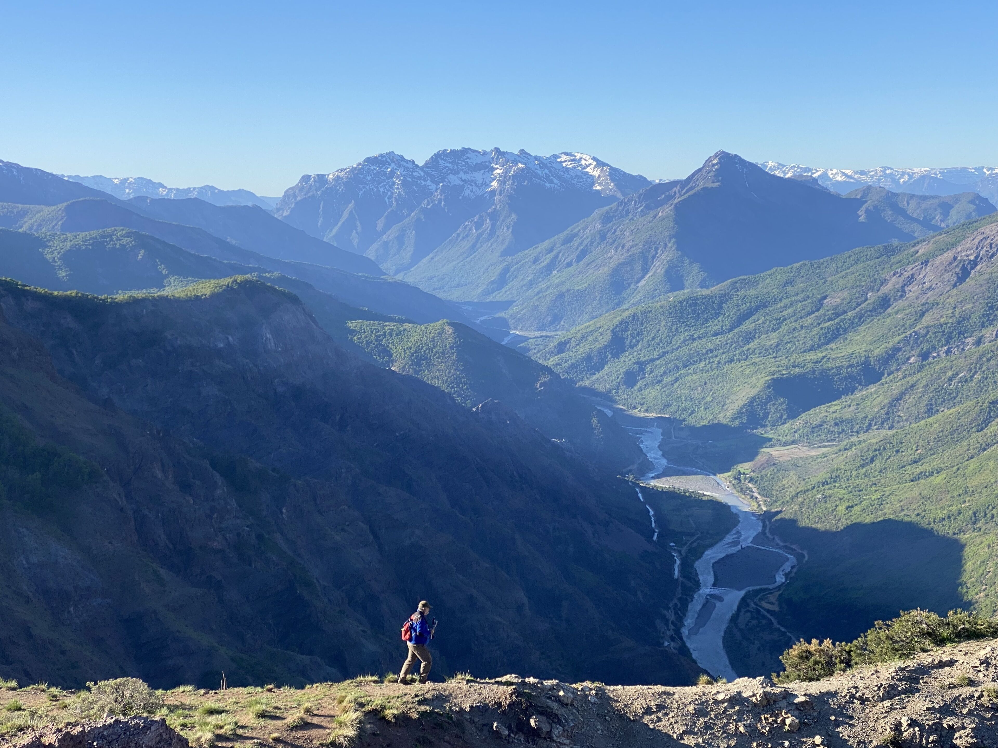 A hiker on a mountain trail with a river and mountains in the background.