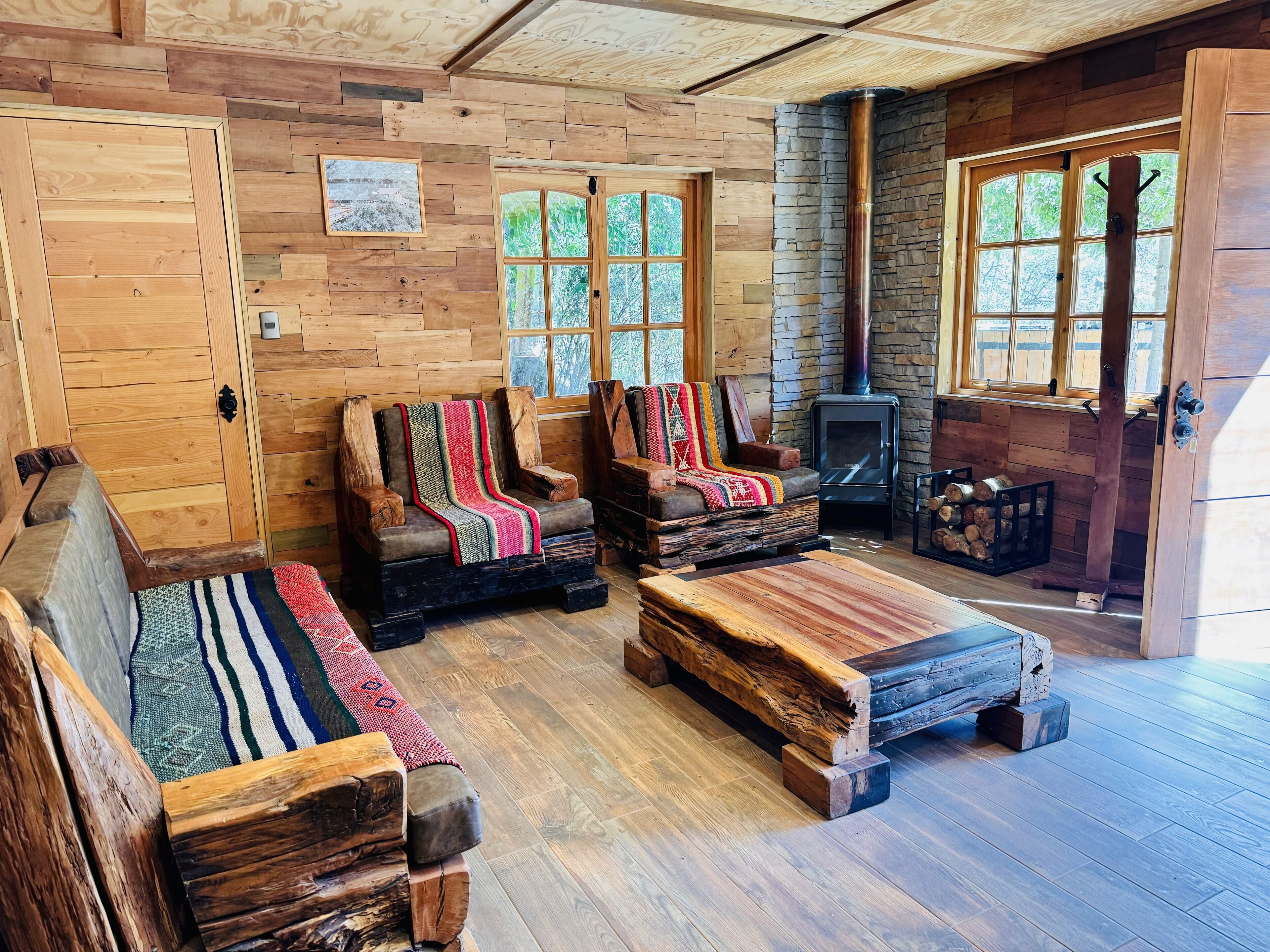 A rustic cabin interior with wooden walls, a stone fireplace, and a cozy seating area with benches and a sofa.
