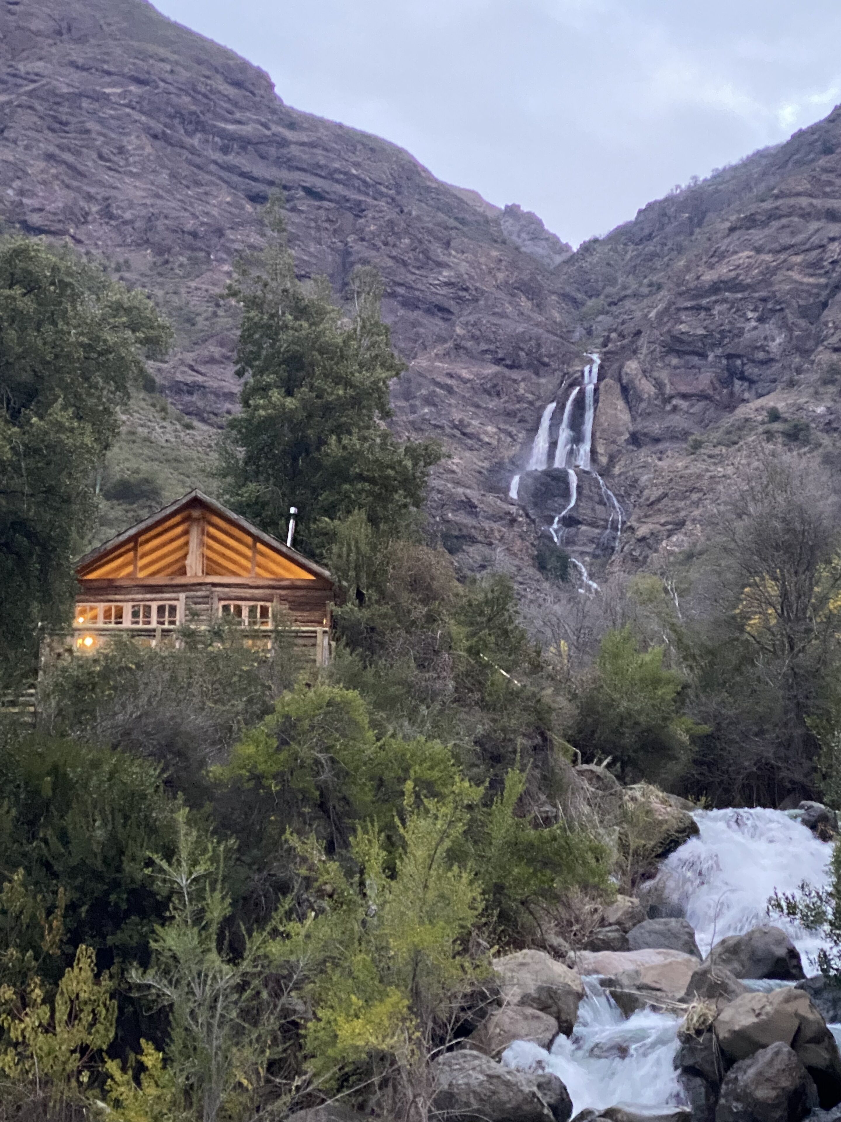 Una cabaña rústica con fachada de madera y chimenea, rodeada de un bosque denso y una cascada al fondo.