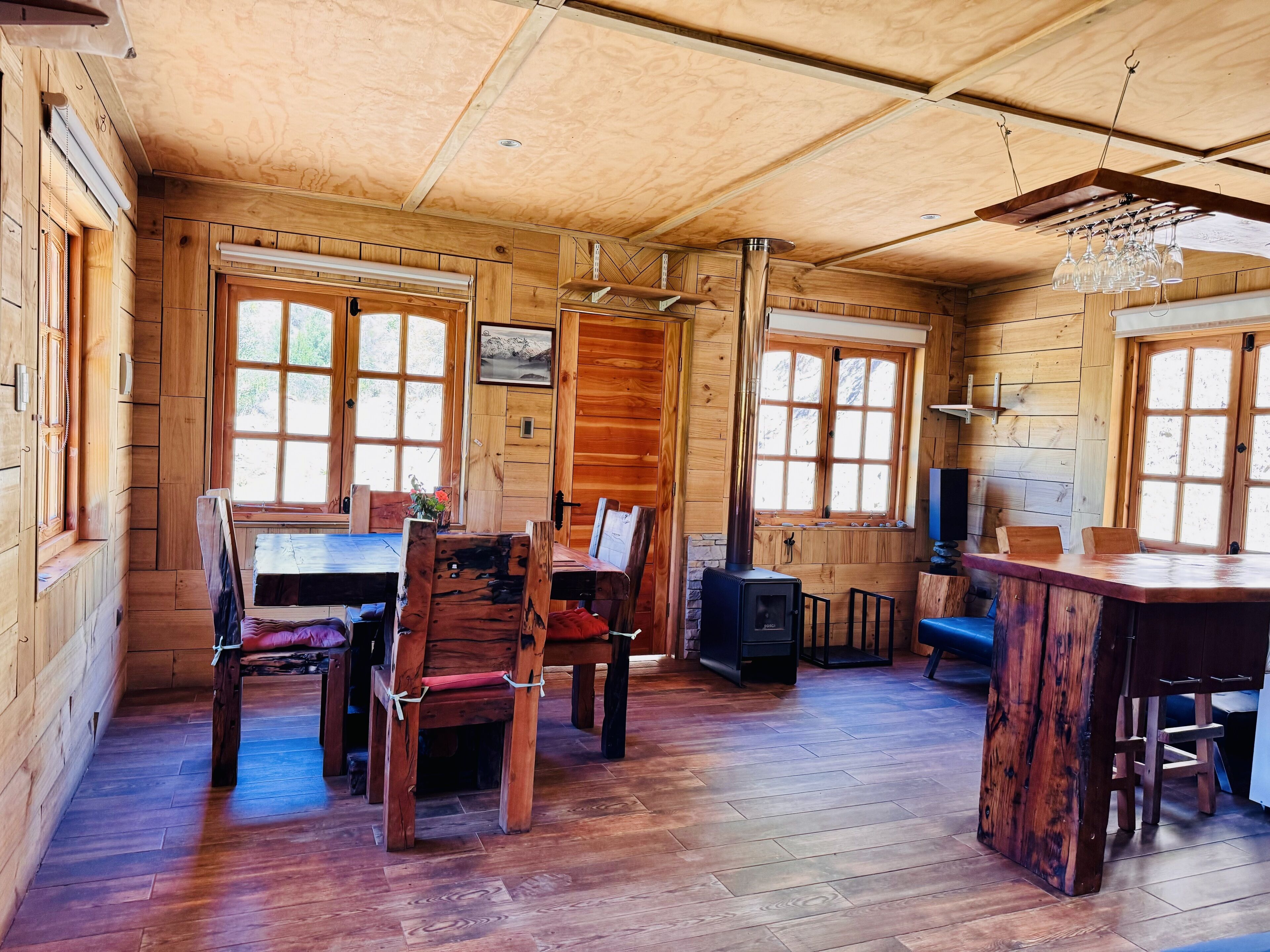 A wooden cabin interior with a dining area, a fireplace, and large windows.