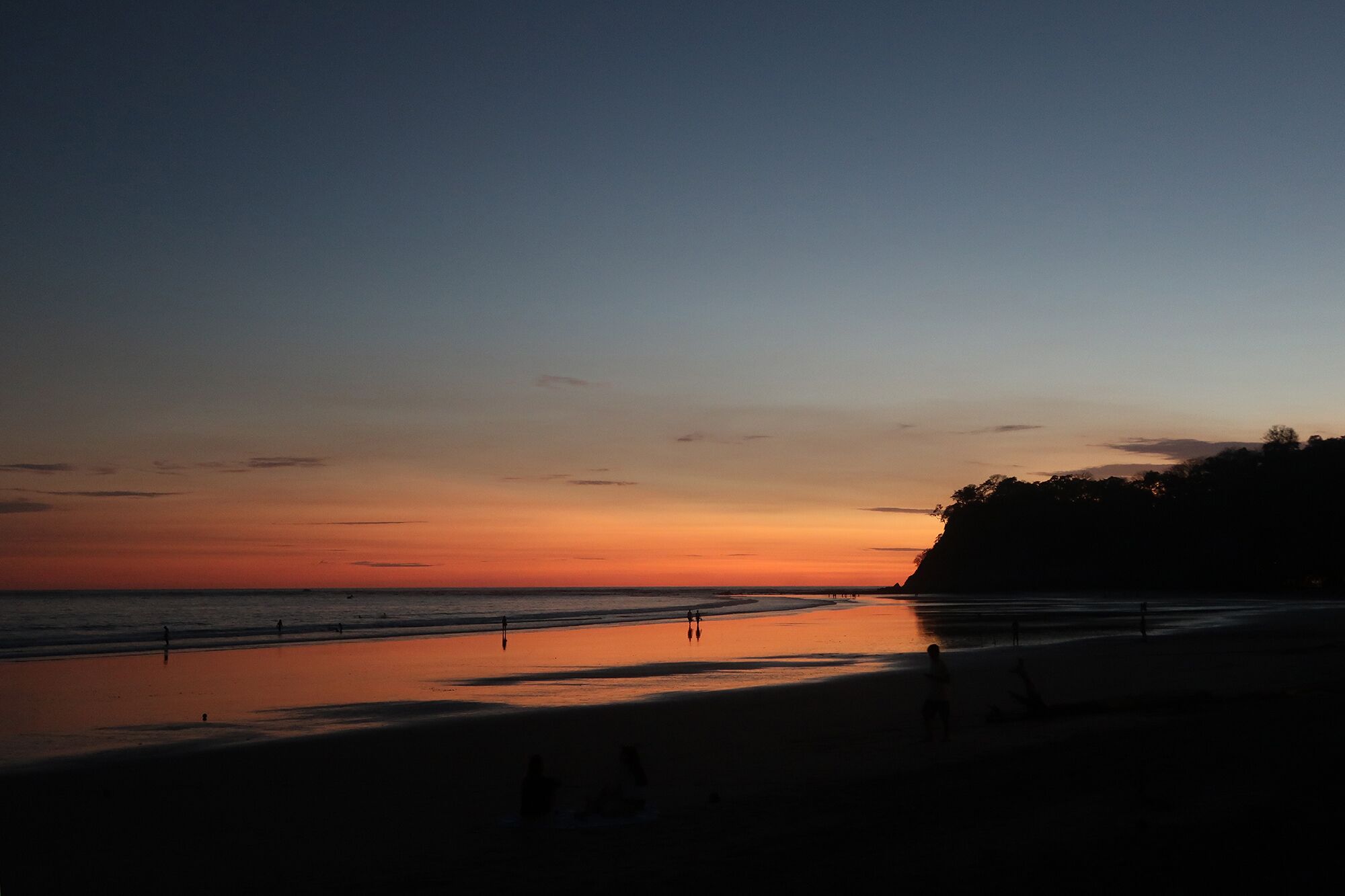 Praia ao pôr do sol com silhuetas de pessoas e céu limpo.