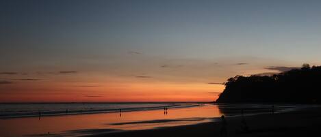Praia ao pôr do sol com silhuetas de pessoas e céu limpo.