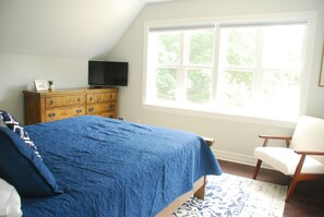 A bedroom with a large bed, a dresser, a television, and a window with a view of trees.