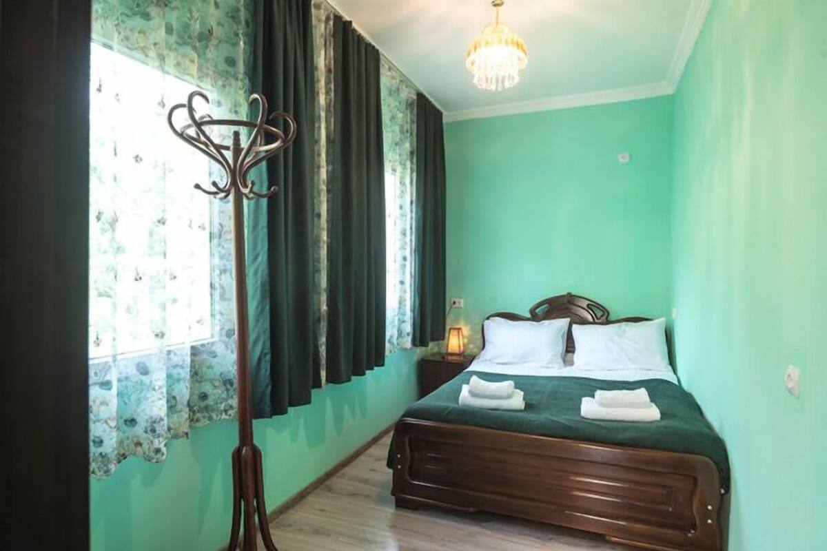 A room with a bed, a nightstand, a lamp, a window with curtains, and a chandelier.