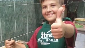 Une personne portant un tablier vert orné d’un logo, tenant un couteau et faisant un geste de pouce levé.