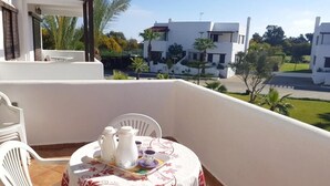 En balkon med et bord dækket til the, hvide stole og udsigt over moderne bygninger og grønne områder.