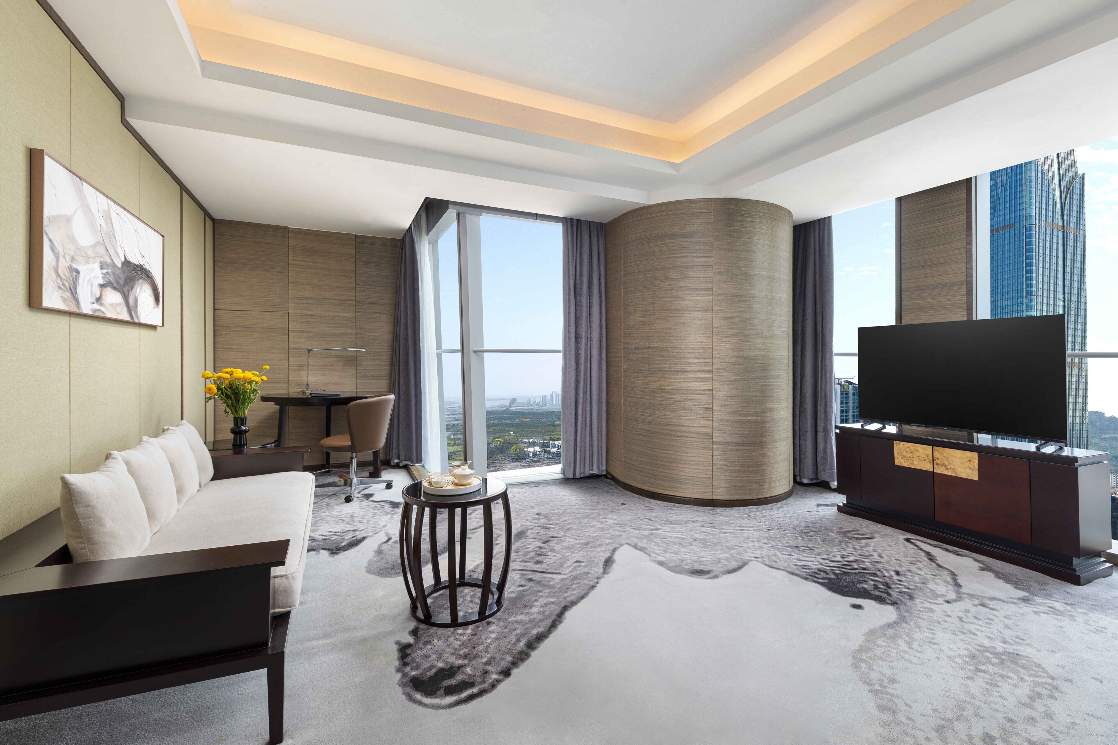 A modern hotel room with a sofa, a flat-screen TV, a dining table, and a large window offering a city view.