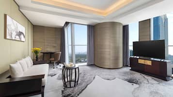 A modern hotel room with a sofa, a flat-screen TV, a dining table, and a large window offering a city view.