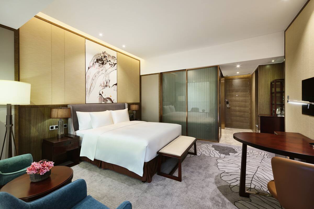 Habitación de hotel con cama, mesita de noche, lámpara, silla, banco y televisor.