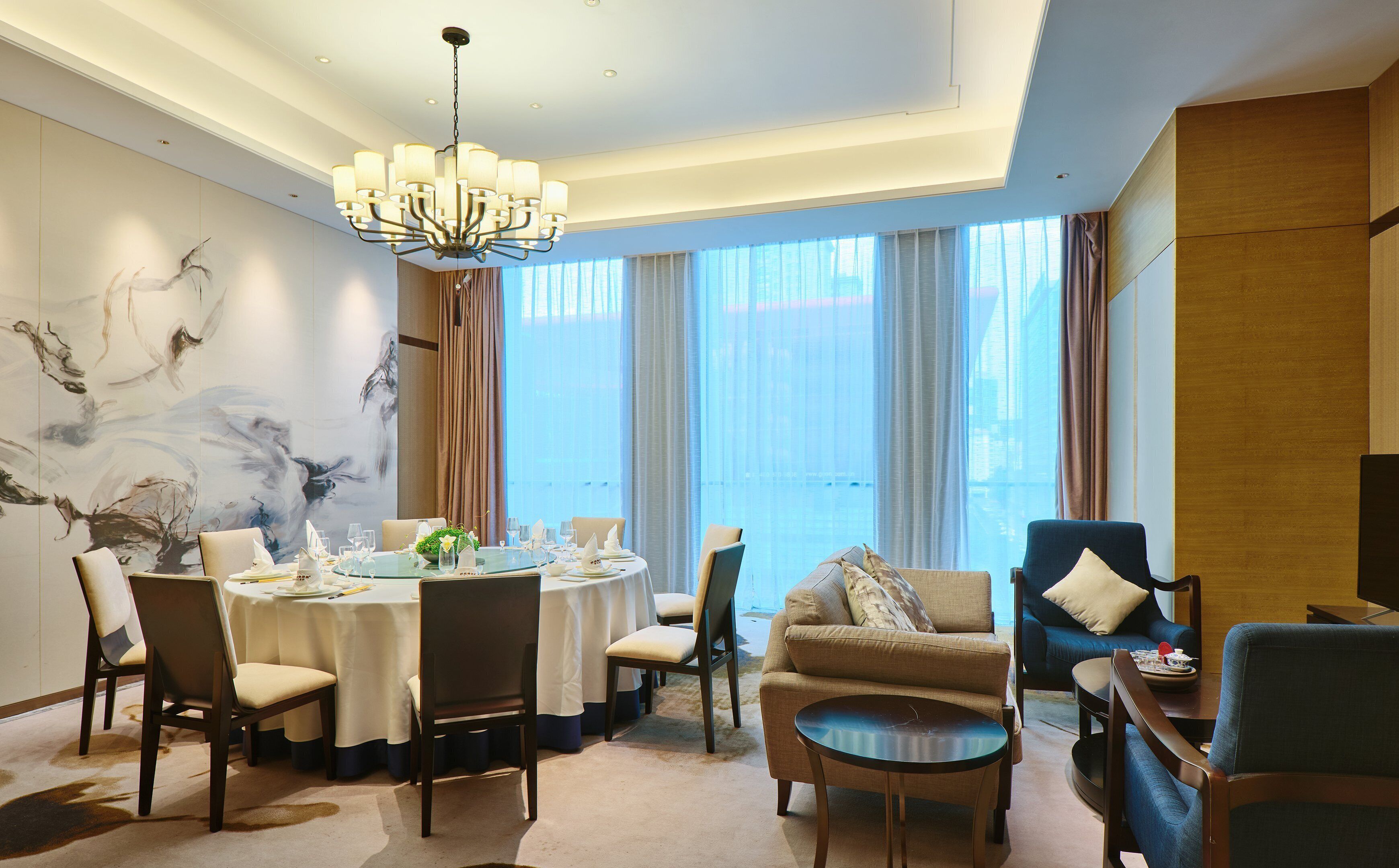 A dining area with a round table set for six, surrounded by chairs, a chandelier, and a large window with curtains.