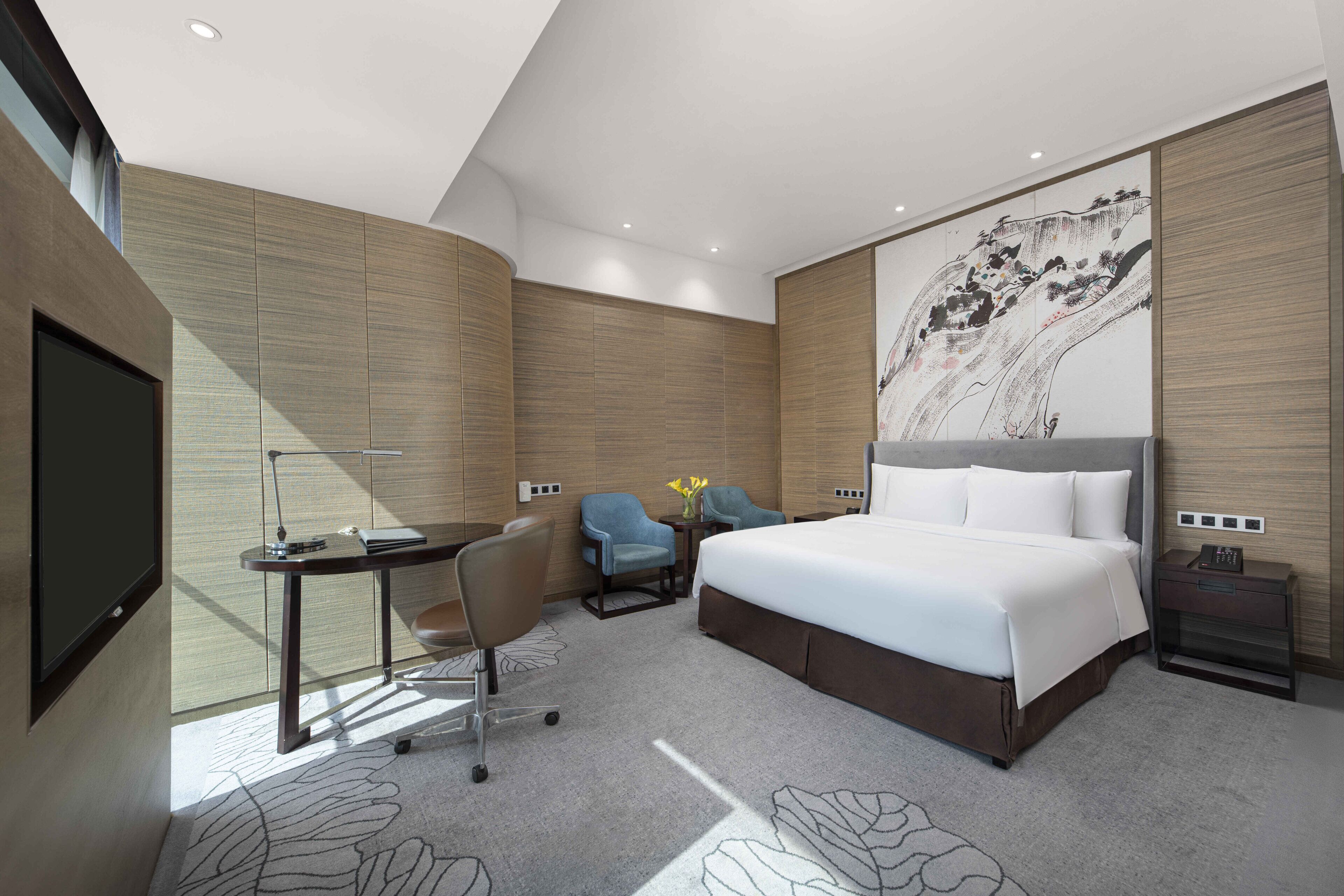 A modern hotel room with a large bed, a desk, two chairs, a TV, and a wall-mounted artwork.