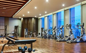 Un gimnasio moderno con varios equipos de ejercicio, incluyendo cintas de correr y máquinas de pesas, y amplias ventanas que ofrecen vistas de la ciudad.