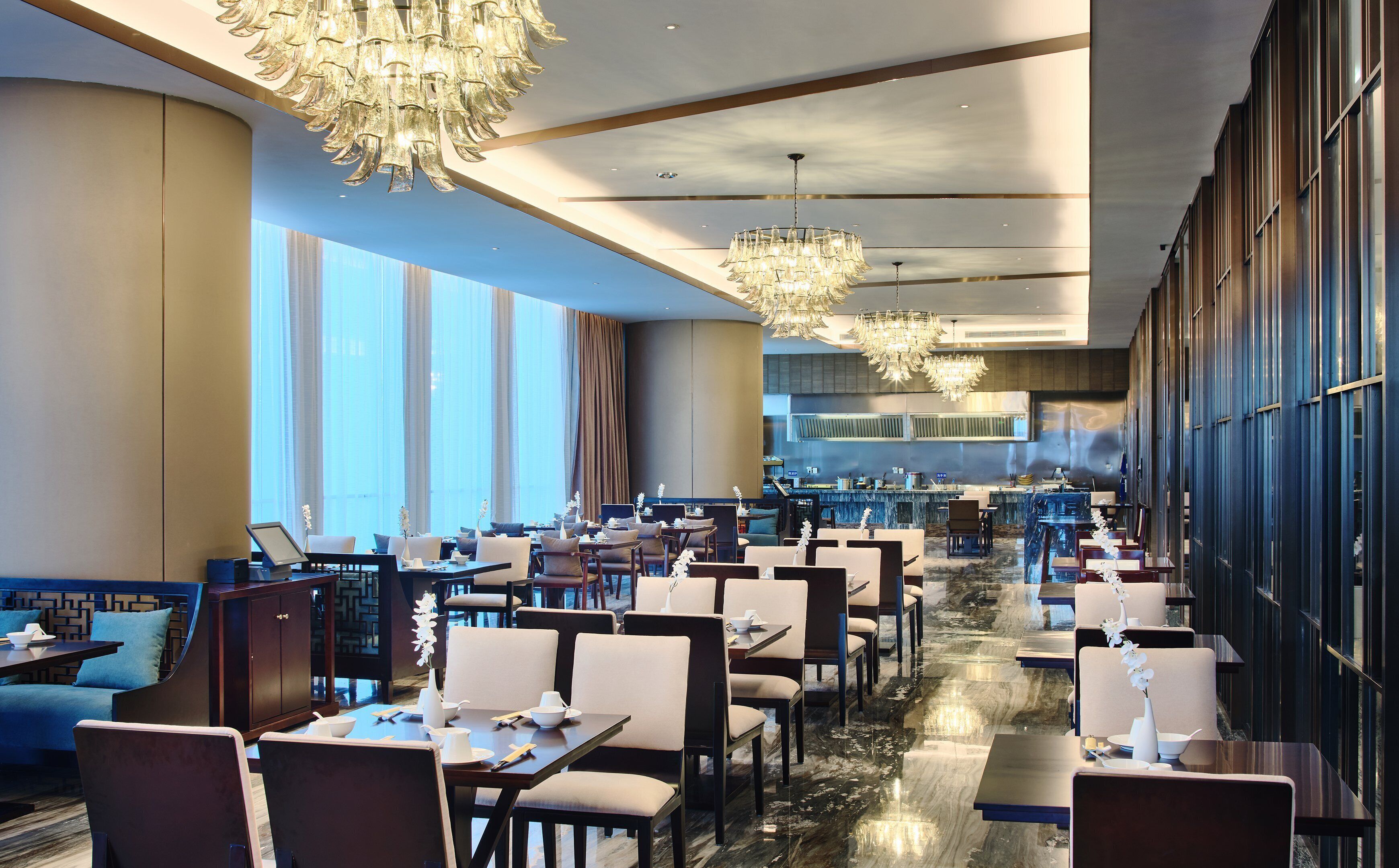Un restaurant spacieux, doté de nombreuses tables et chaises, de grandes fenêtres et d’un lustre moderne.