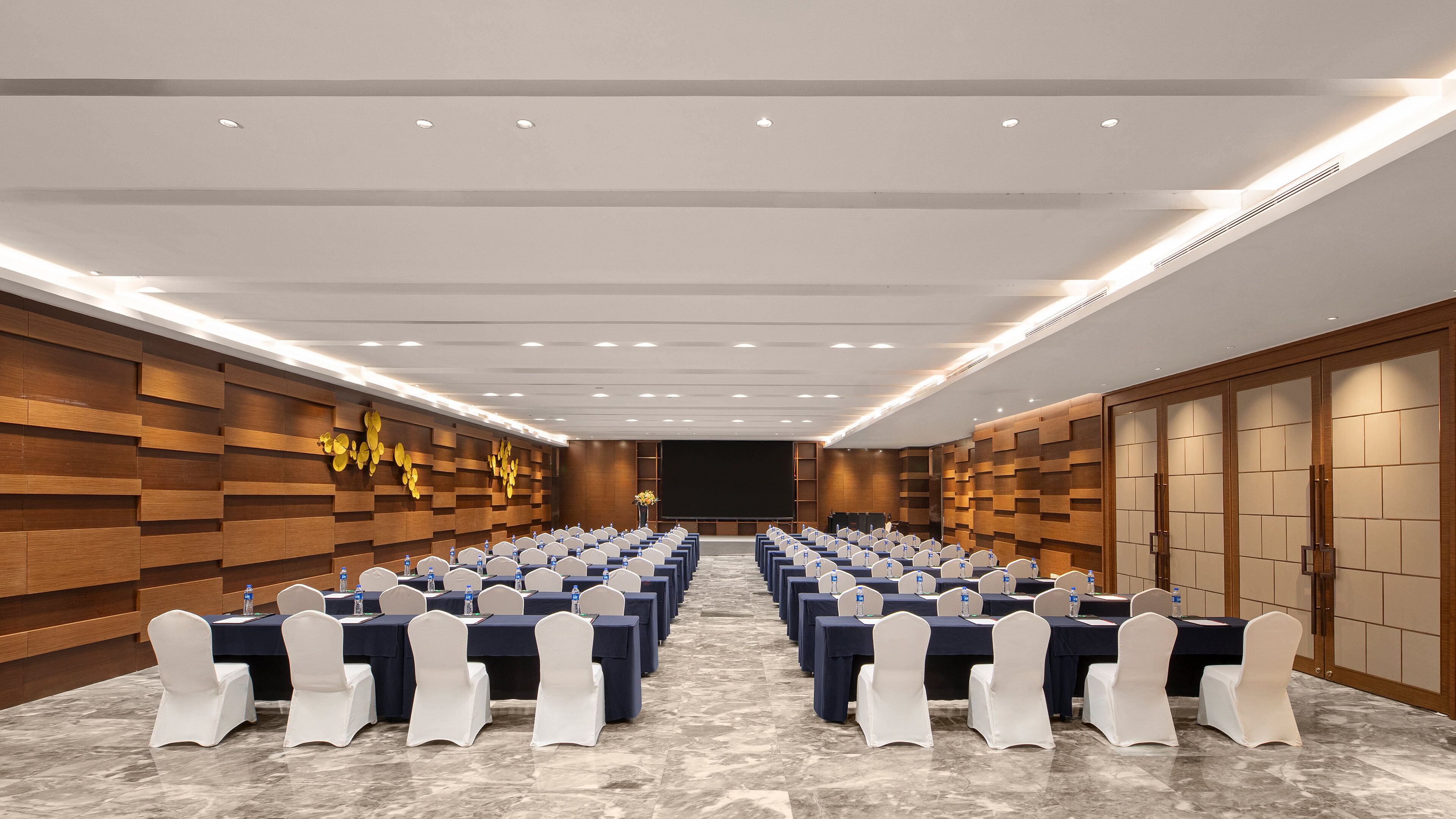 A conference room with rows of tables and chairs, a stage, and wooden panel walls.