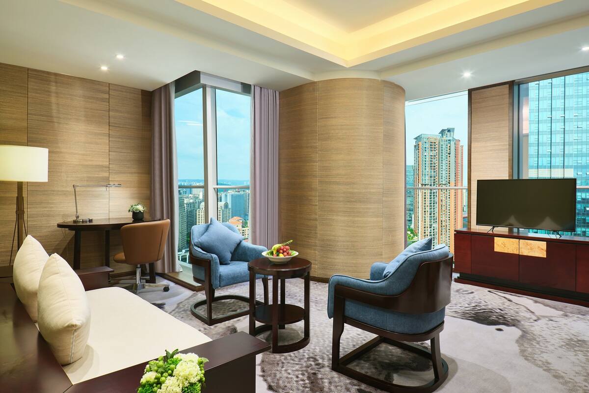 Una habitación de hotel moderna con un ventanal grande, un televisor de pantalla plana, un sofá, dos sillones, una mesita con frutero y un escritorio con lámpara.