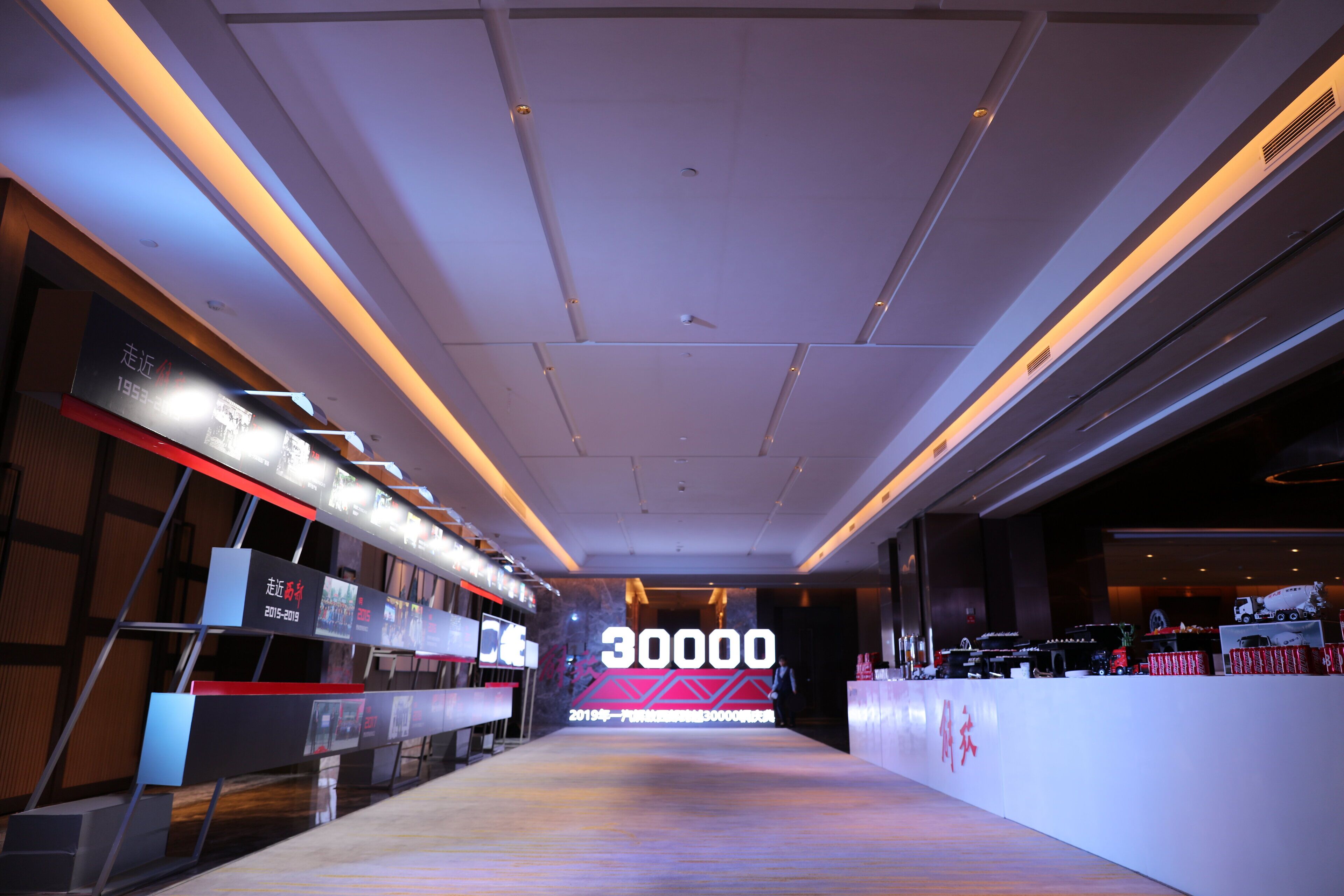 A modern hotel lobby with a long reception desk, a large digital display, and a high ceiling with recessed lighting.