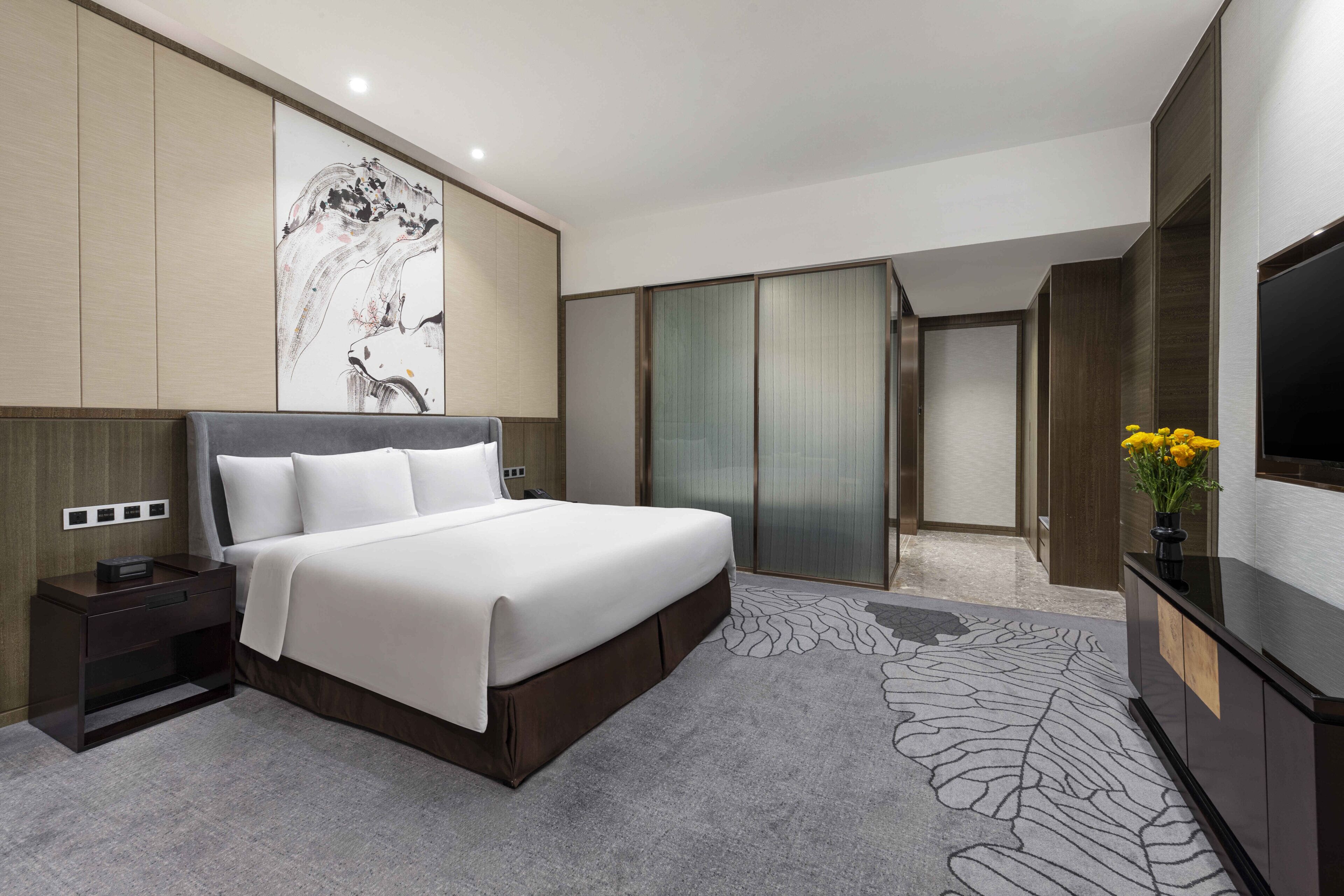 A modern hotel room with a large bed, a flat-screen TV, a wardrobe, and a decorative wall piece.