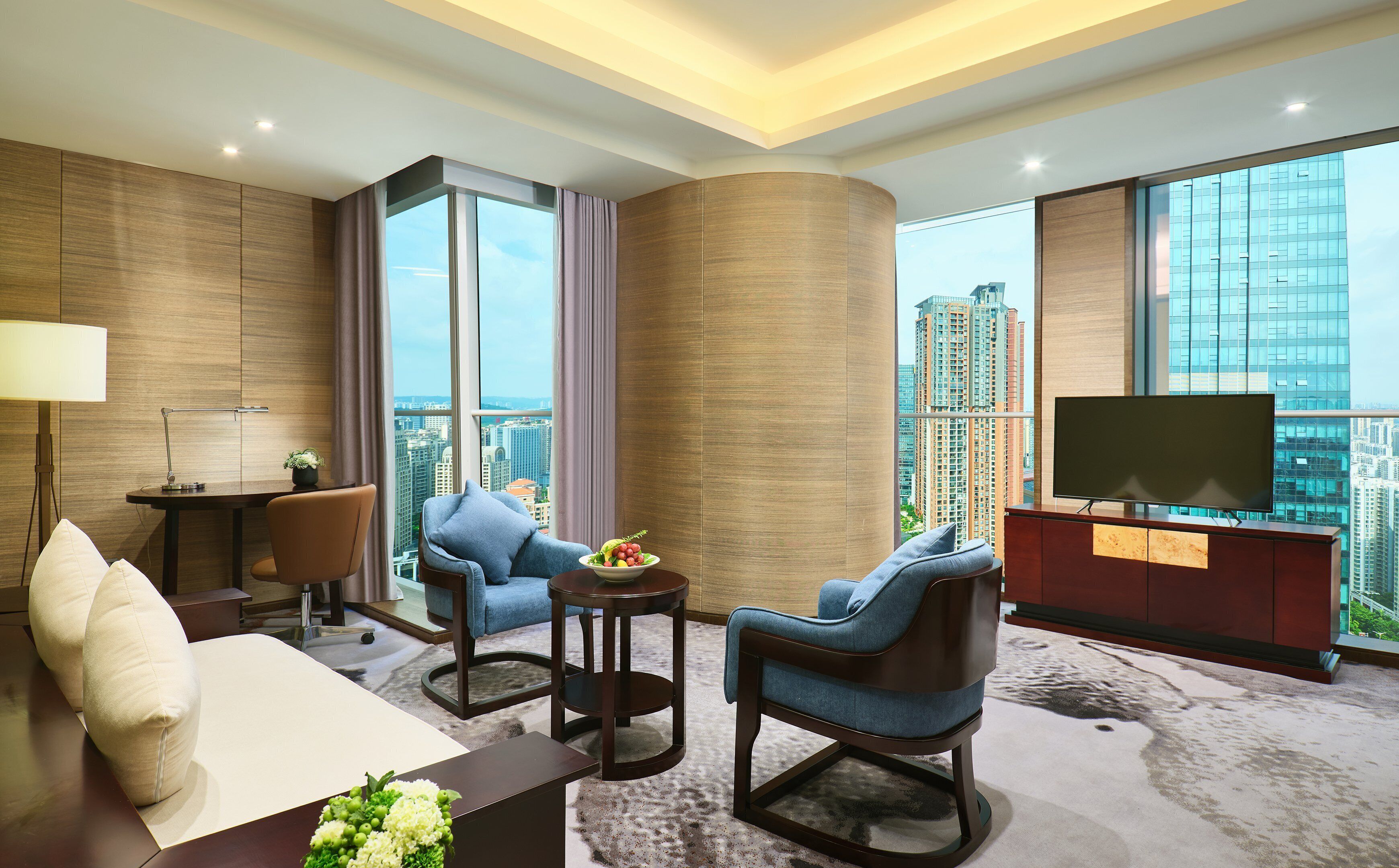 A hotel room with a large window, a flat-screen TV, a sofa, two armchairs, a small table with a bowl of fruit, and a desk with a lamp.