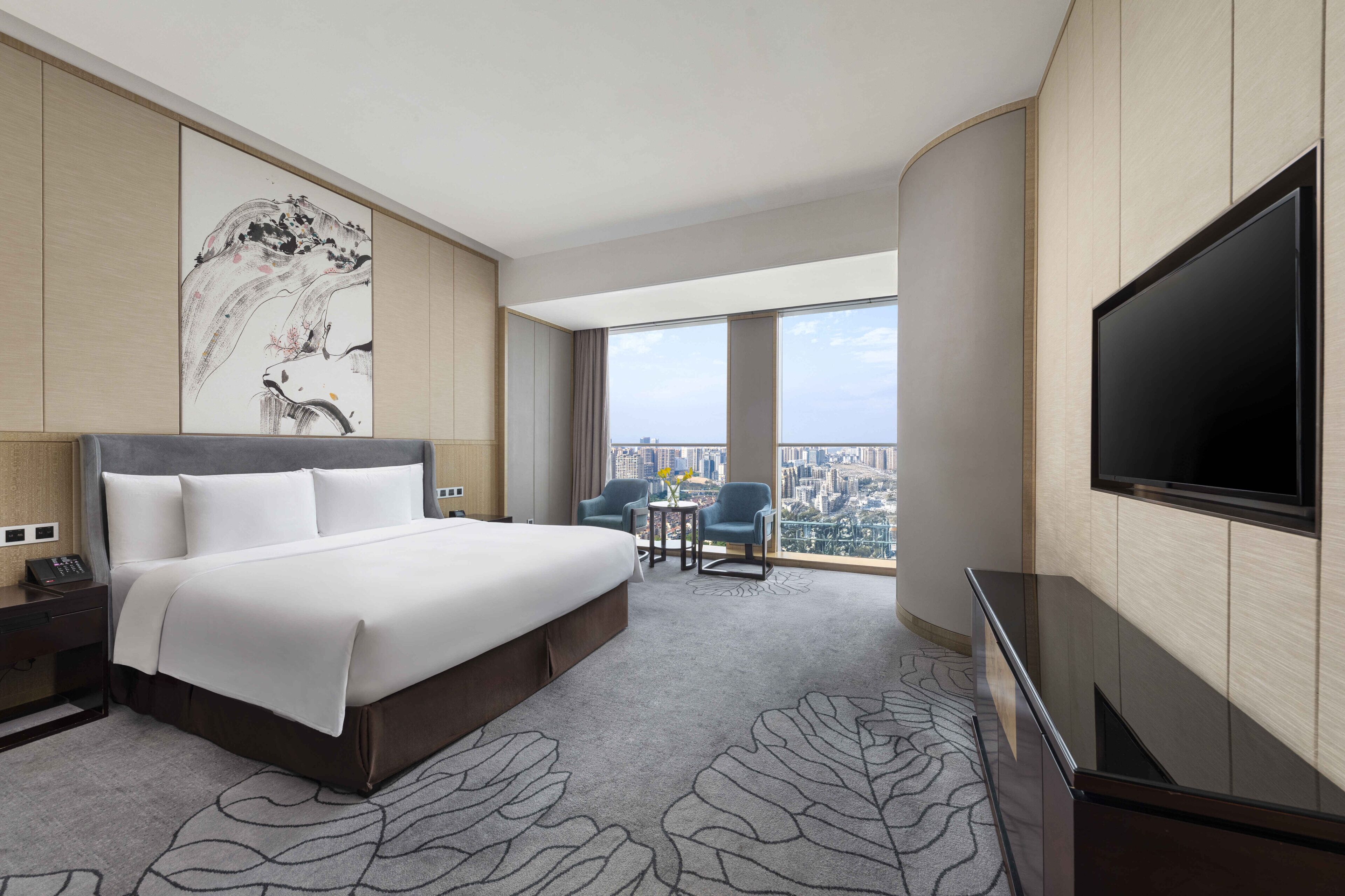 A hotel room with a large bed, a TV, a desk with a phone, a chair, and a view of the city.