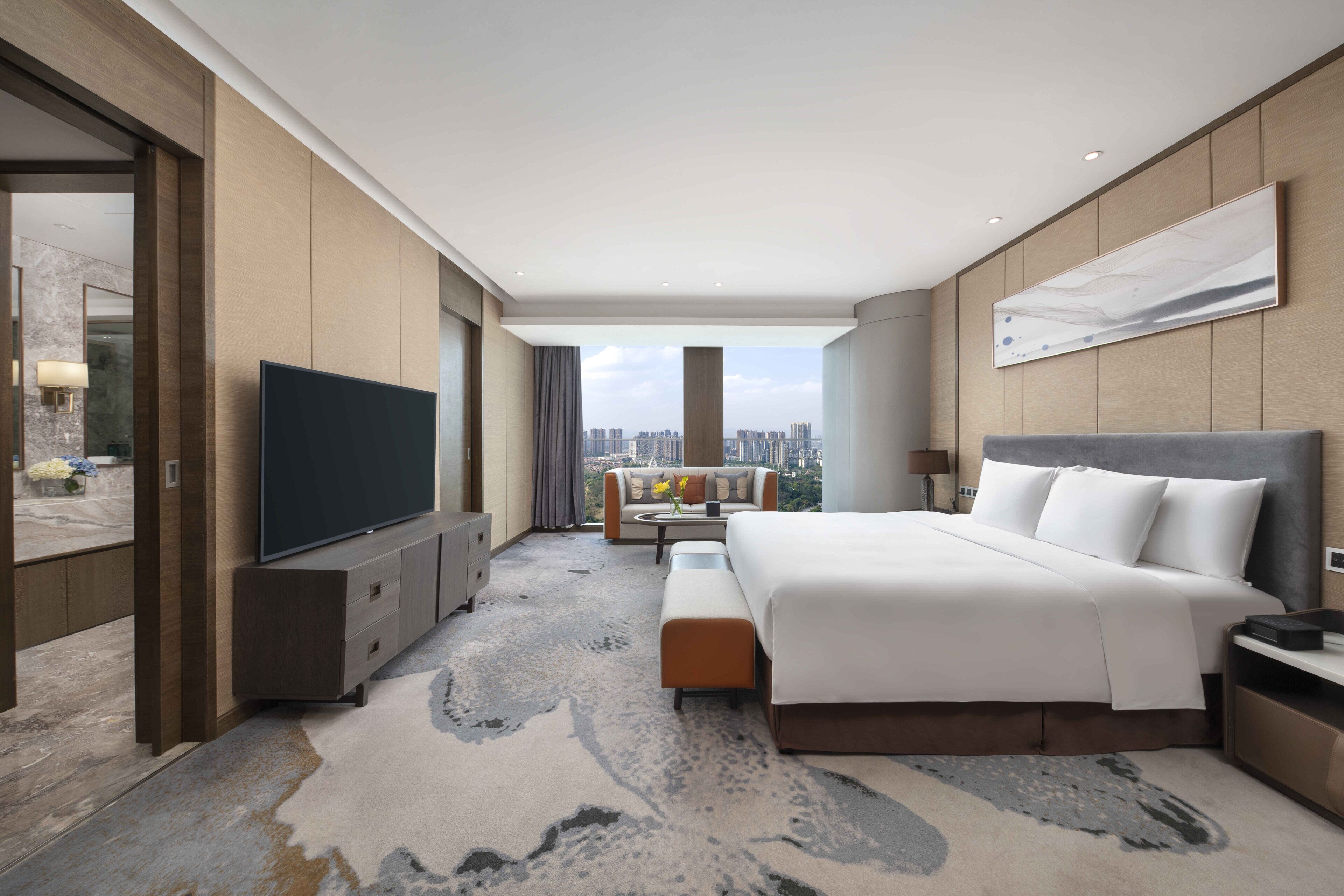 A modern hotel room with a large bed, a flat-screen TV, a sofa, and a city view.