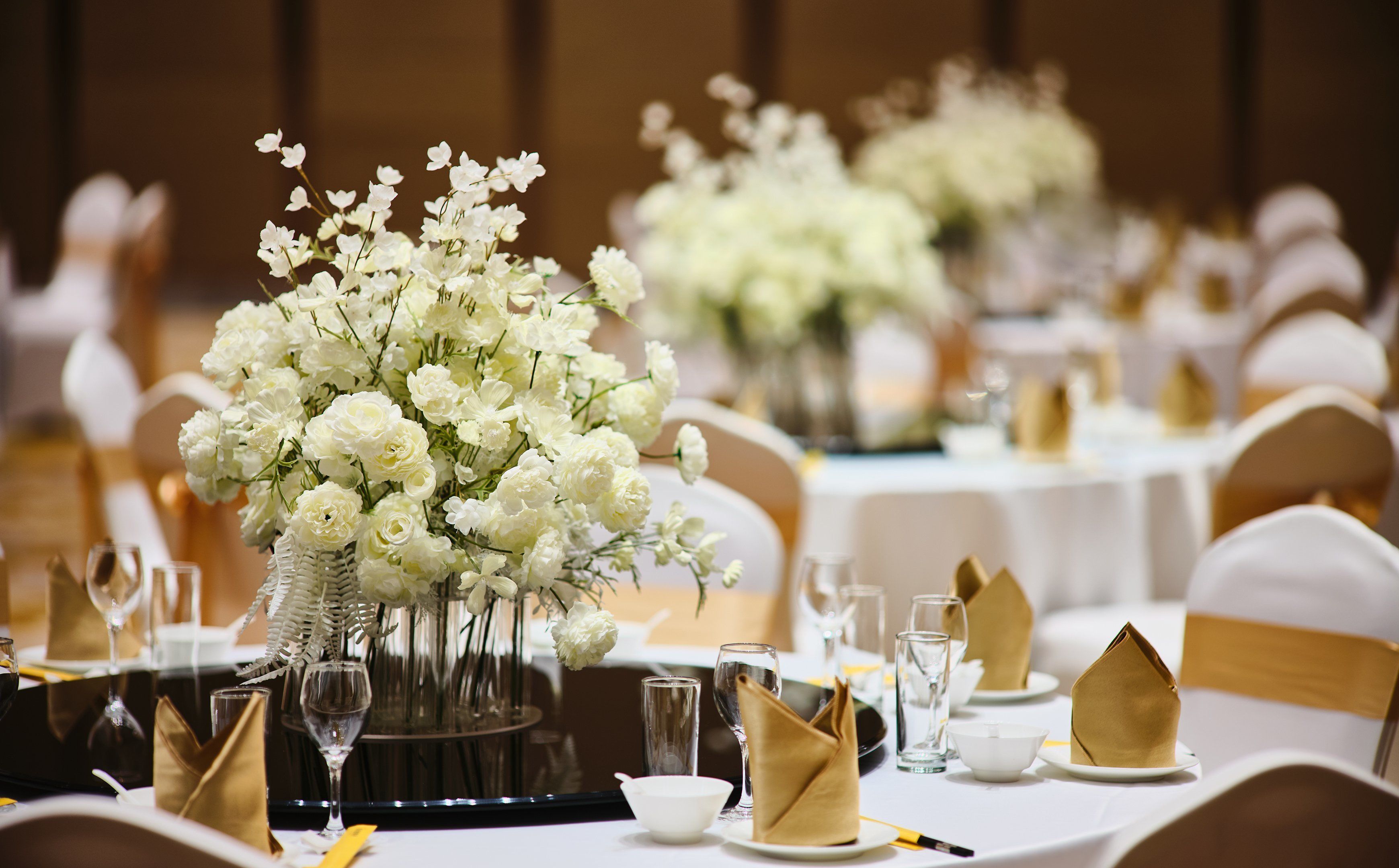 Salão de festas com mesas redondas preparadas para um evento formal, com centros de mesa florais brancos e guardanapos dourados.