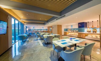 Un comedor moderno de hotel con mesas y sillas, una zona de cocina y un amplio ventanal con vistas.