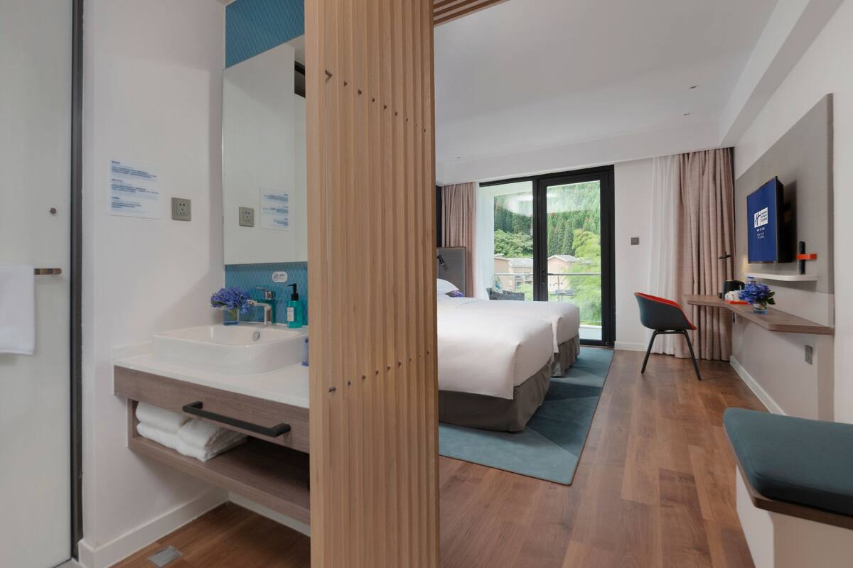 A modern hotel room with a large bed, a bathroom with a sink and mirror, a desk with a chair, and a TV on the wall.