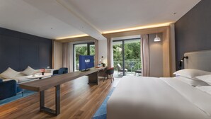 A modern hotel room with a large bed, a desk with a computer, a seating area with a sofa and chairs, and large windows offering a view of greenery. - Holiday Inn Express E'Mei Qiliping by IHG (Meishan)
