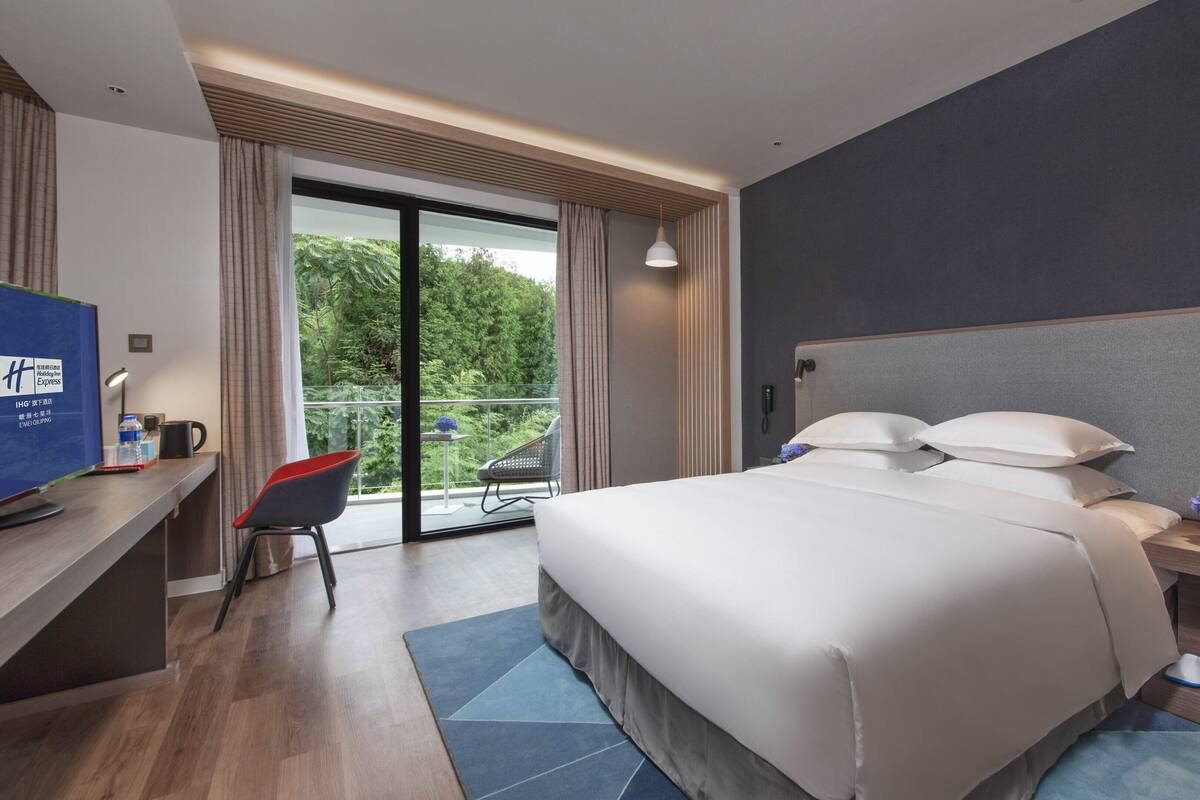 A modern hotel room with a large bed, a desk with a computer, a chair, a small table with a lamp, and a balcony with a view of trees.