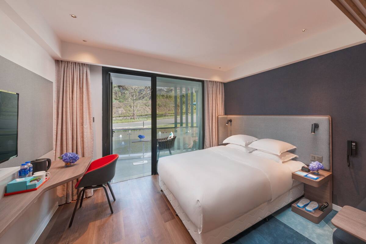 A modern hotel room with a large bed, a desk with a chair, a TV, and a view of the outdoors.